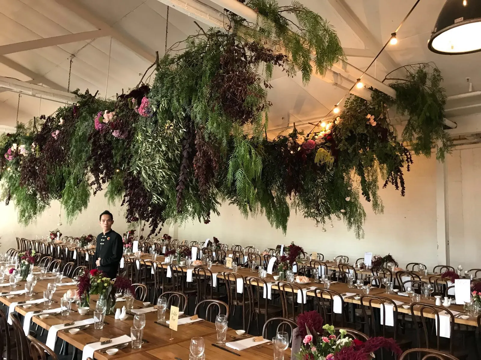 A hanging floral ceiling installation of greenery and burgundy blooms over long wedding reception tables